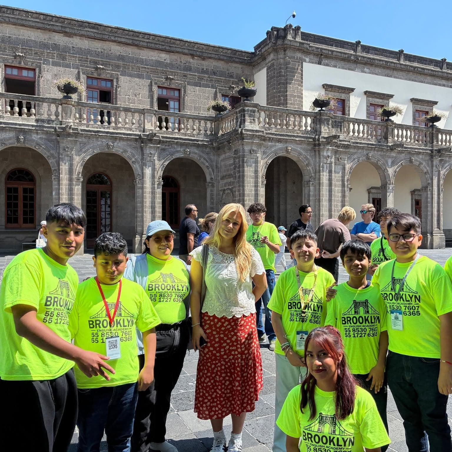 Grupo de alumnos de Brooklyn con playera amarilla junto a una visitante extranjera, frente a un edificio histórico